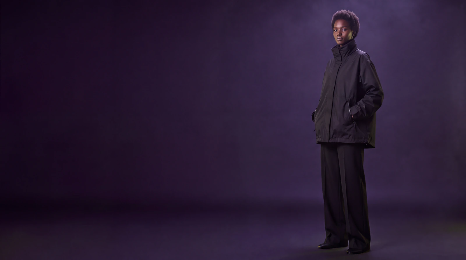 A woman in an oversized black technical shell jacket and wide black trousers, standing in a dark violet studio